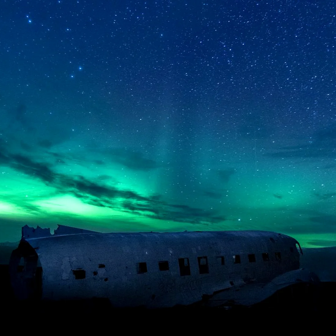 A stripped aircraft fuselage lying on black sand under a green aurora and a dense starfield